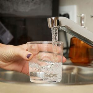 A hand with painted nails holding a clear glass under a modern kitchen faucet as filered water flows into the glass, with a blurred kitchen background.